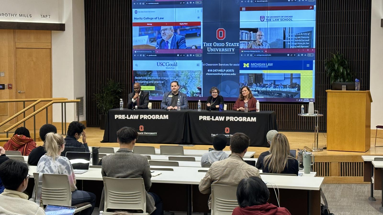 Panel of speakers in front of an audience, video monitors in background showing websites